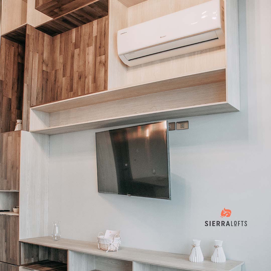 A modern living area is presented with a wall-mounted television and air conditioning unit above. Shelves, made of light and dark wood, are arranged for storage and display. A small decorative basket is placed on the shelf, enhancing the overall functionality.