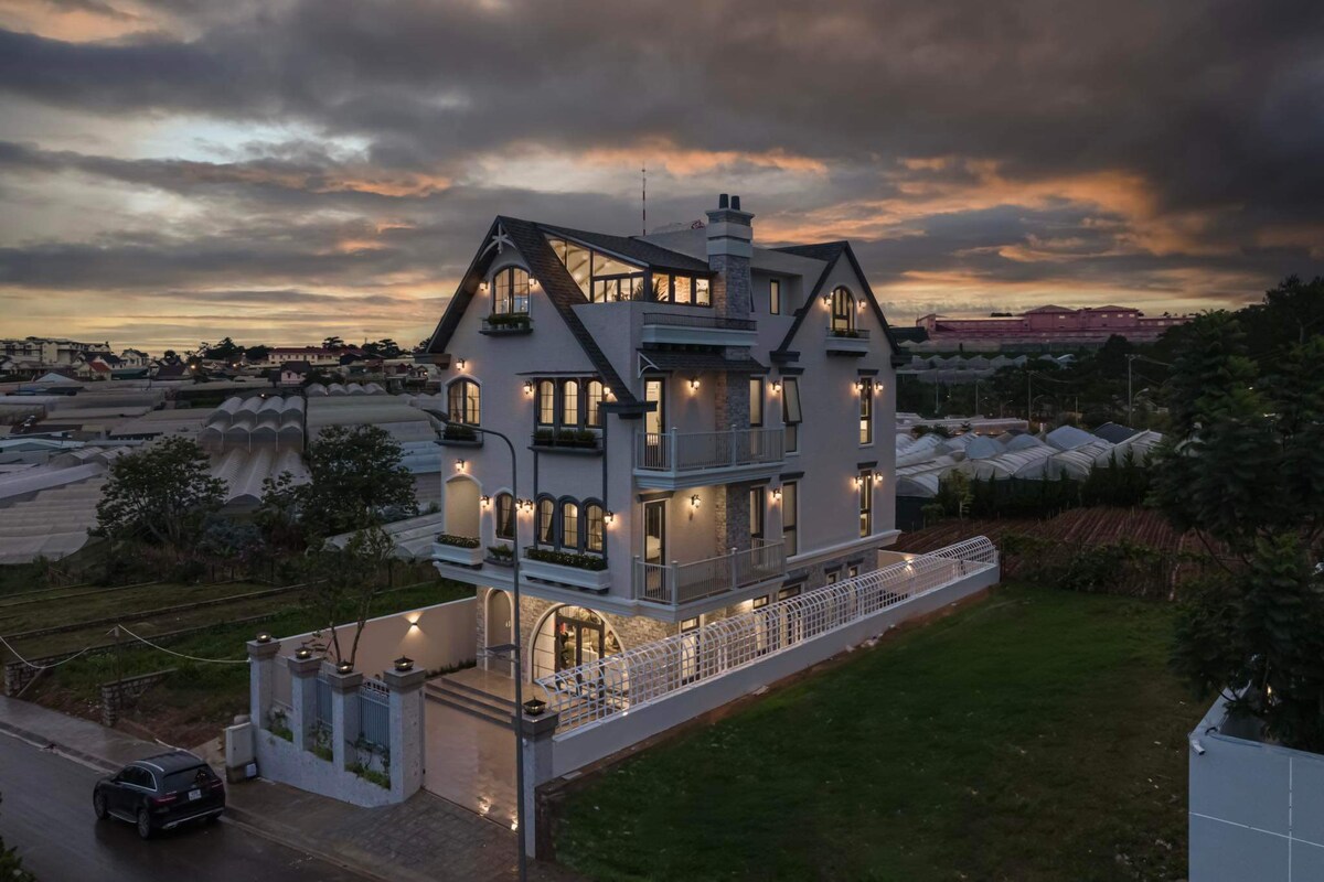 A three-story villa showcases elegant architecture, featuring multiple windows and a sloped roof. Soft lighting outlines the exterior, complementing the colorful twilight sky. The surrounding green space is complemented by a paved driveway, creating an inviting entrance.