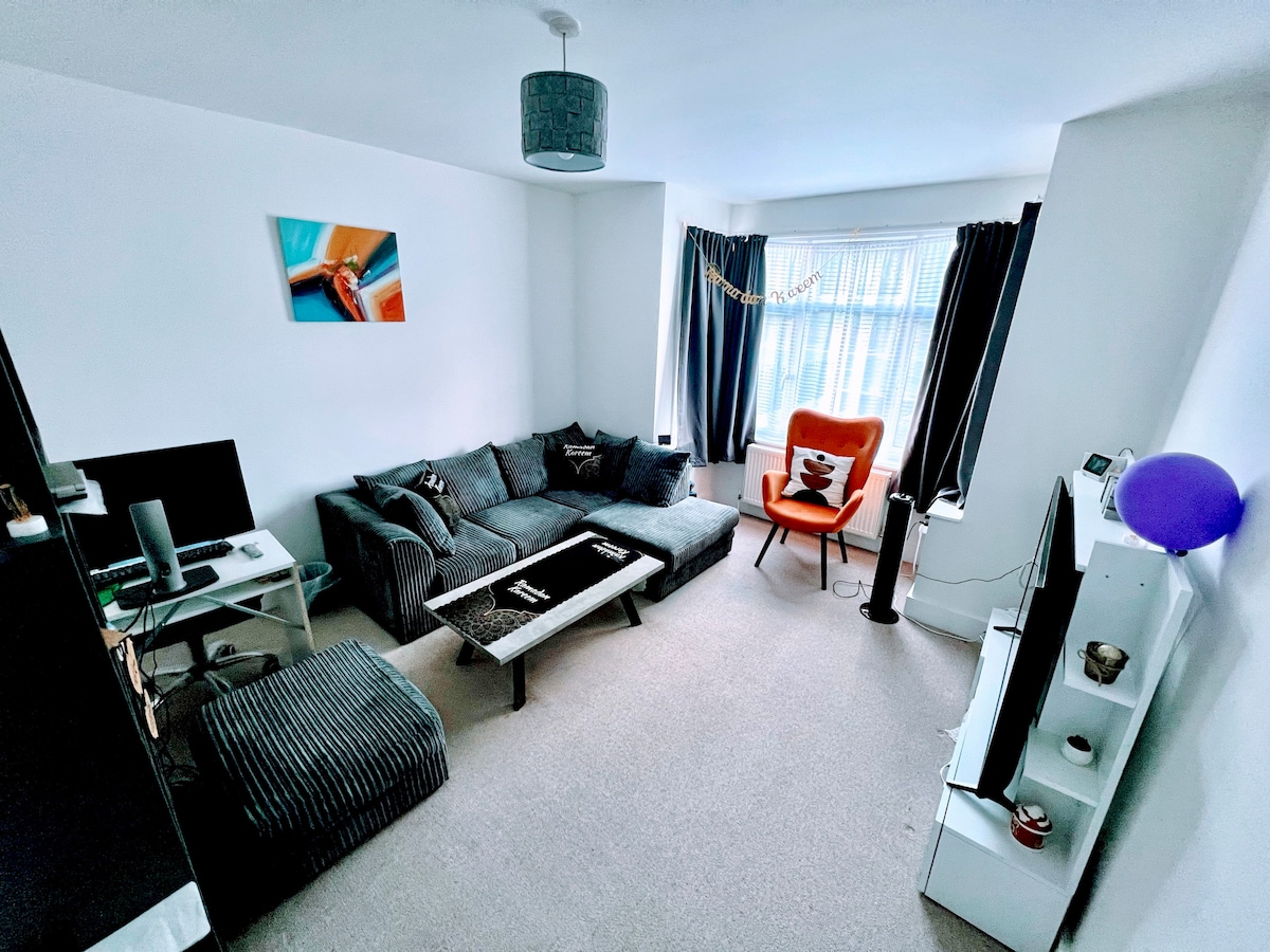 The living area features a cozy grey sectional sofa arranged around a low coffee table. A bright orange chair adds a pop of color. Natural light enters through the large window, which is framed by dark curtains. A flat-screen TV is positioned on a modern white console.