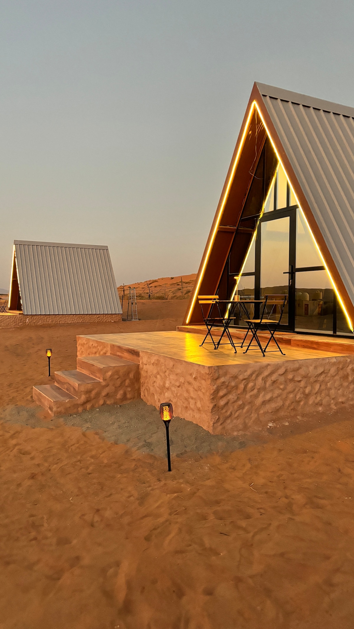 A unique triangular structure is set against a desert backdrop, featuring a wooden deck with a small table and two chairs. Soft lighting frames the edges, highlighting the architecture. The sandy landscape surrounds the cabin, contributing to a serene atmosphere.