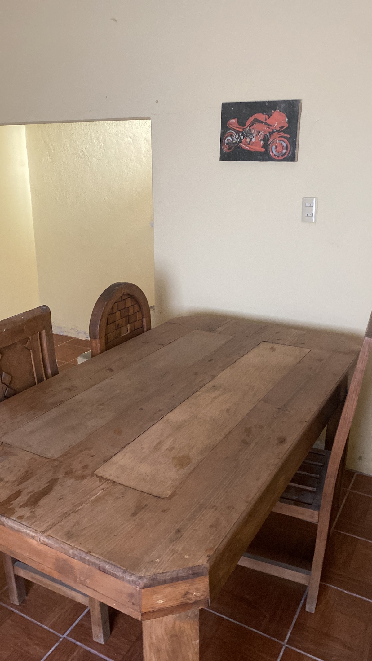 A wooden dining table, showing a rustic finish, is surrounded by matching chairs. A small artwork depicting a motorcycle hangs on the wall nearby. The space is brightened by natural light, enhancing the simplicity and warmth of the dining area.