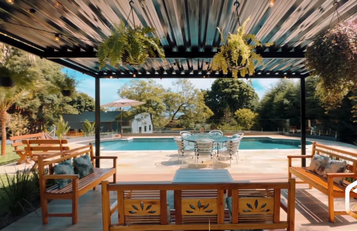 A shaded seating area is created beneath a metal pergola, featuring wooden benches and a coffee table. Potted ferns hang from above. In the background, a large pool is visible, surrounded by lush greenery and clear blue skies.