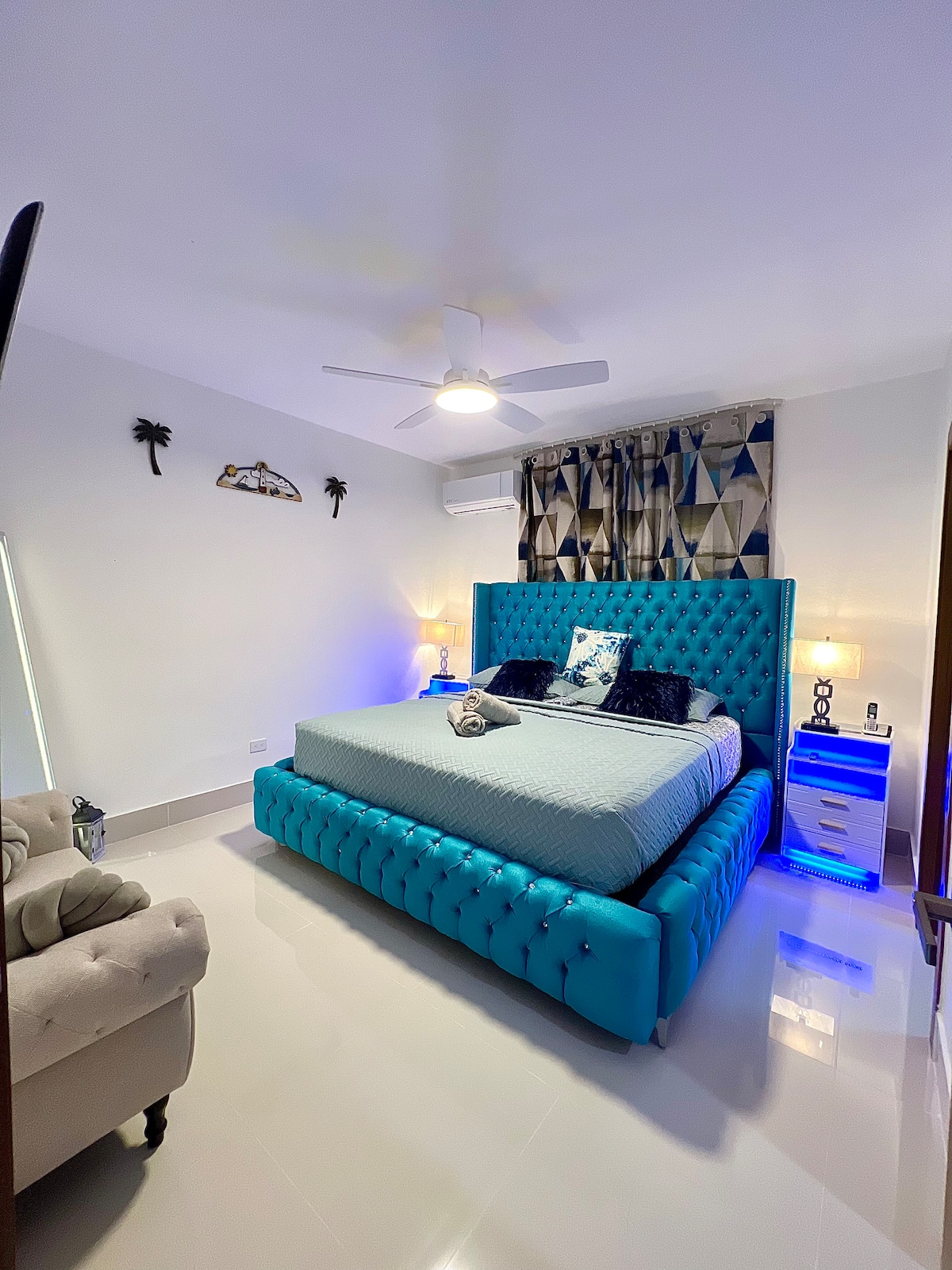 A spacious bedroom features a turquoise tufted bed with decorative pillows and a patterned headboard. Soft lighting from bedside tables enhances the room's ambiance. A ceiling fan and air conditioning unit are present, with light-colored walls and a window adorned by patterned curtains.