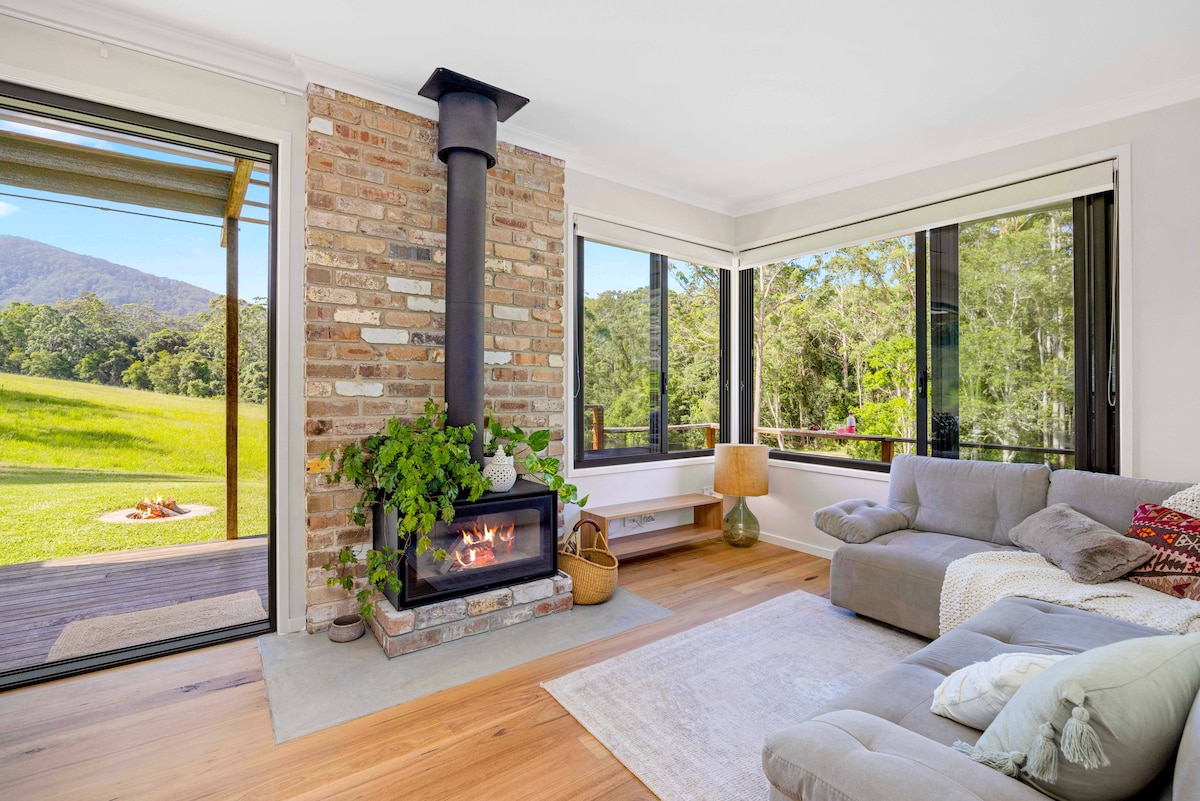 A modern living area features a brick fireplace as a focal point, surrounded by expansive windows that frame views of the lush outdoor landscape. A cozy sofa with decorative cushions and a soft area rug completes the inviting space, allowing natural light to fill the room.
