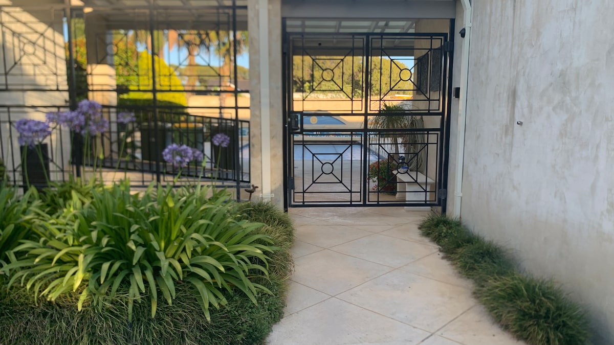 A gated entrance features decorative black ironwork framing a view of the pool area in the distance. Lush greenery lines the pathway, complemented by purple flowers and neatly trimmed grass, creating a welcoming approach to the property.