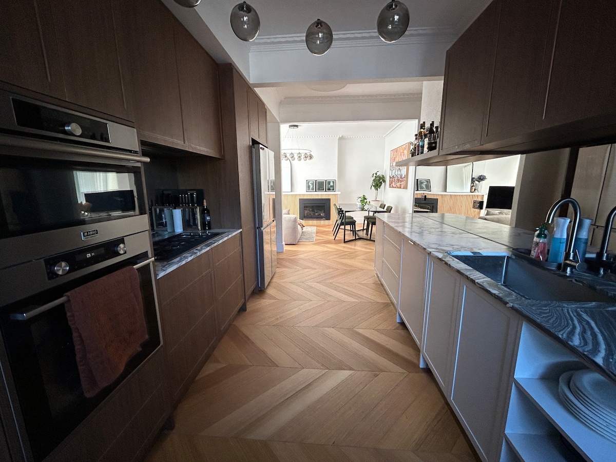 A modern kitchen is showcased with sleek cabinetry and marble countertops. Features include a gas stove, oven, and coffee maker. The open layout connects to the living area visible in the background, enhancing the sense of space. Natural light fills the area through large windows.