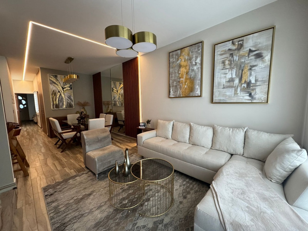 A spacious living area features a light-colored sectional sofa paired with a round coffee table. Two large abstract paintings adorn the walls, while a rug adds texture to the wood flooring. Natural light fills the space through a nearby doorway.
