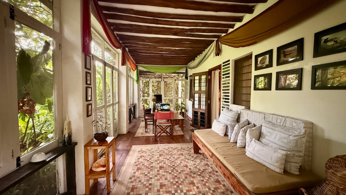 An inviting open living area features a wooden ceiling with exposed beams. Natural light filters through large windows surrounded by greenery. A comfortable sofa is adorned with patterned cushions, while a dining table is set nearby. Decorative frames line the walls, enhancing the calming atmosphere.