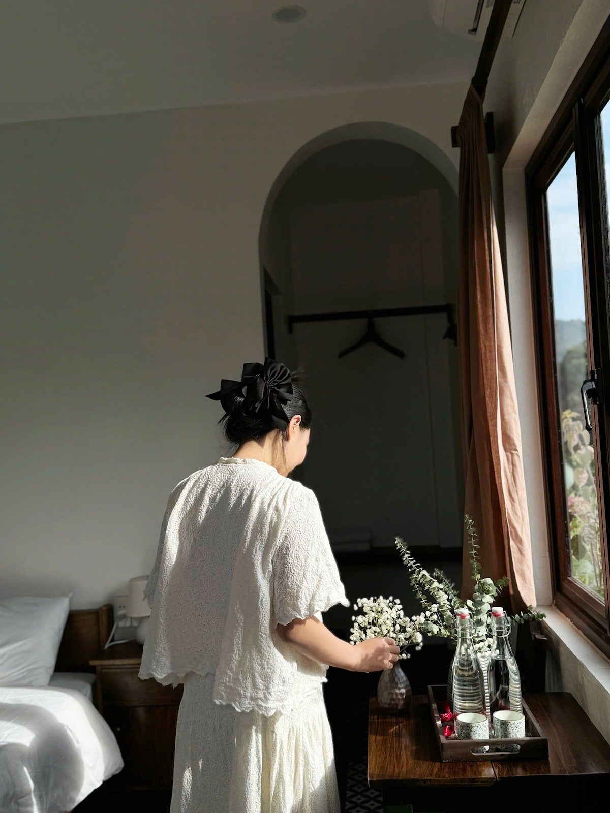 A bright sleeping area features a spacious bed with light bedding. A wooden table holds flower arrangements and glass bottles. Natural light enters through a large window, illuminating the room and creating a serene environment. A closet is seen in the background.