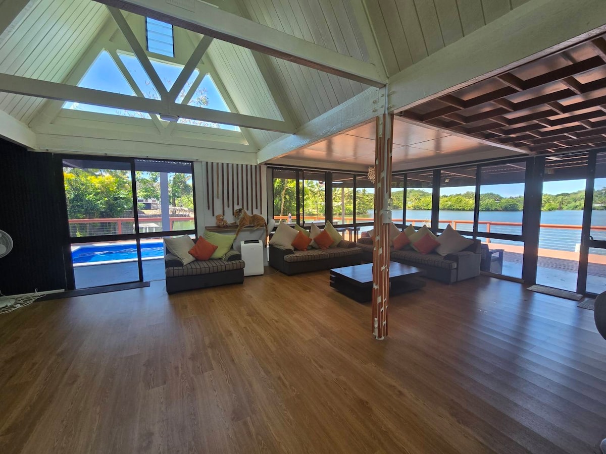 A spacious living area is showcased with large glass windows allowing views of the lake. Wooden flooring complements the light-colored walls and ceiling. Comfortable seating arrangements are presented with decorative cushions. The room is filled with natural light, creating a bright and airy environment.