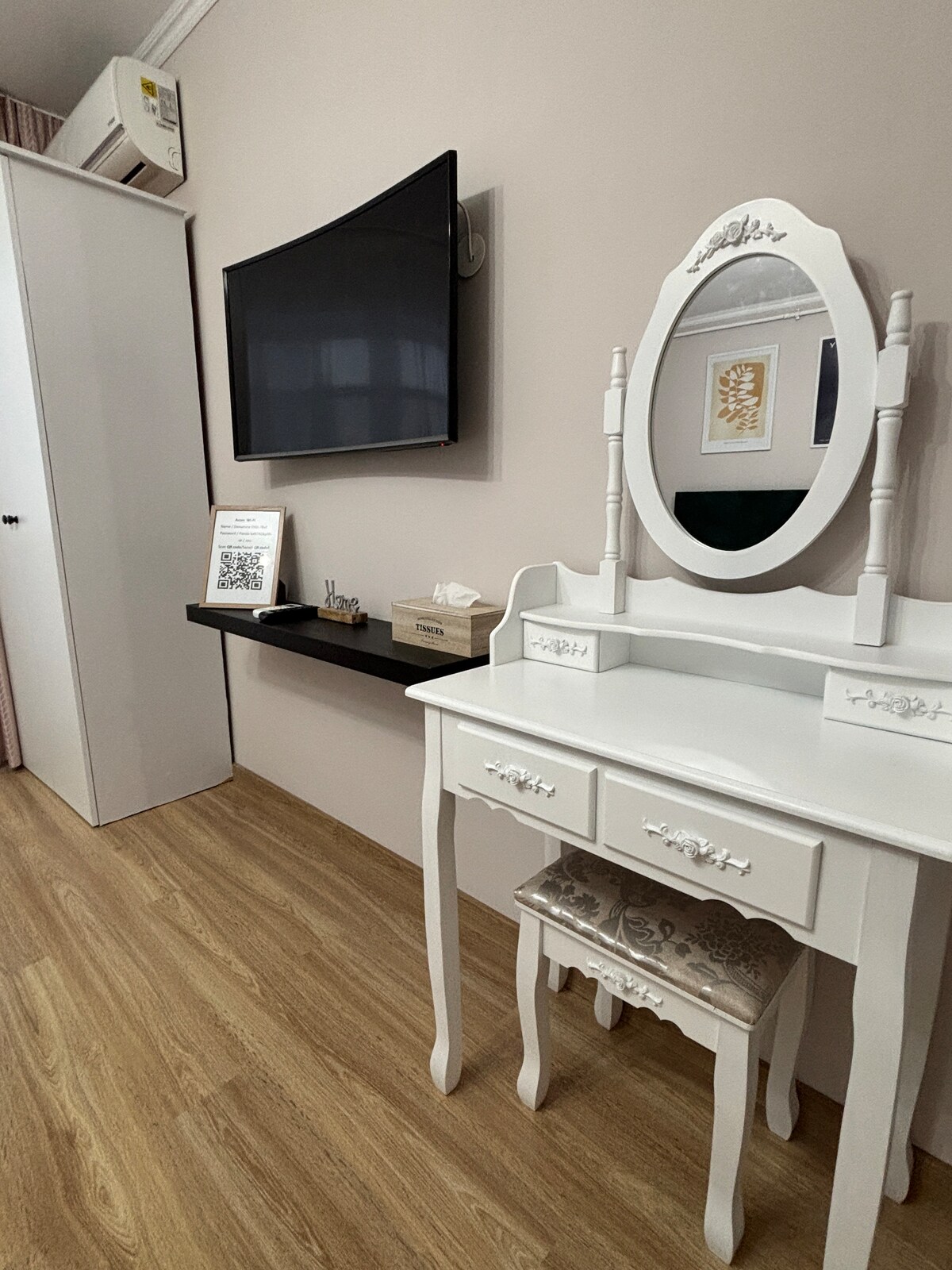 A neatly arranged room features a white vanity with a round mirror and a soft upholstered stool. A wall-mounted television is positioned above a narrow shelf. Light wooden flooring complements the light color scheme, while a closet is visible in the background, enhancing the room's functionality.