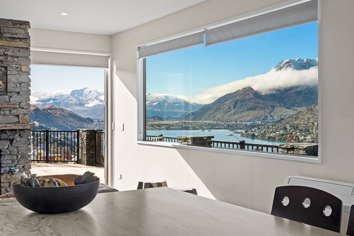 A bright dining area is showcased with a modern table set against a backdrop of large windows. Scenic views of snow-capped mountains and serene waters are captured, enhancing the space with natural light and open vistas.