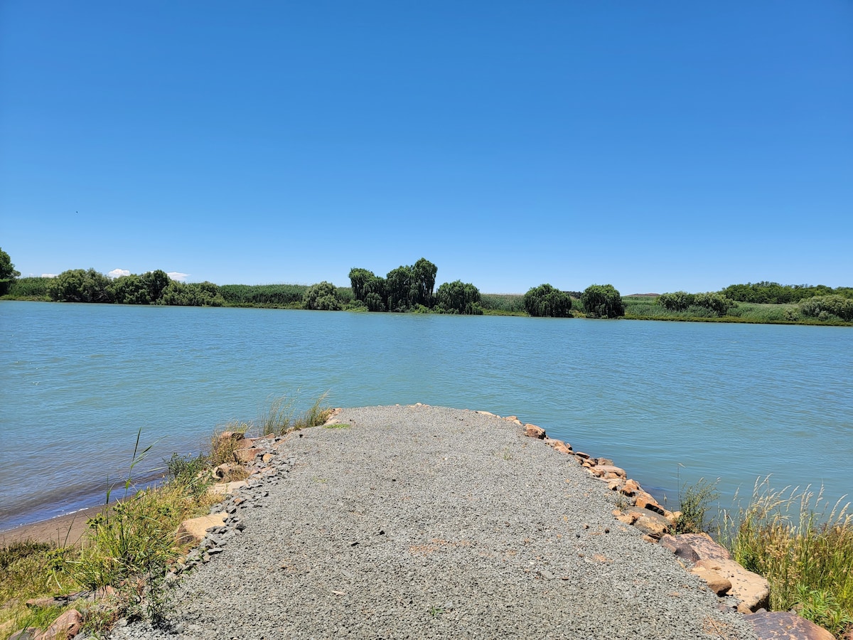 A gravel path leads towards a calm body of water, bordered by lush greenery on the opposite bank. The clear blue sky reflects on the water's surface, creating a serene atmosphere. The area is surrounded by soft grass and occasional bushes.
