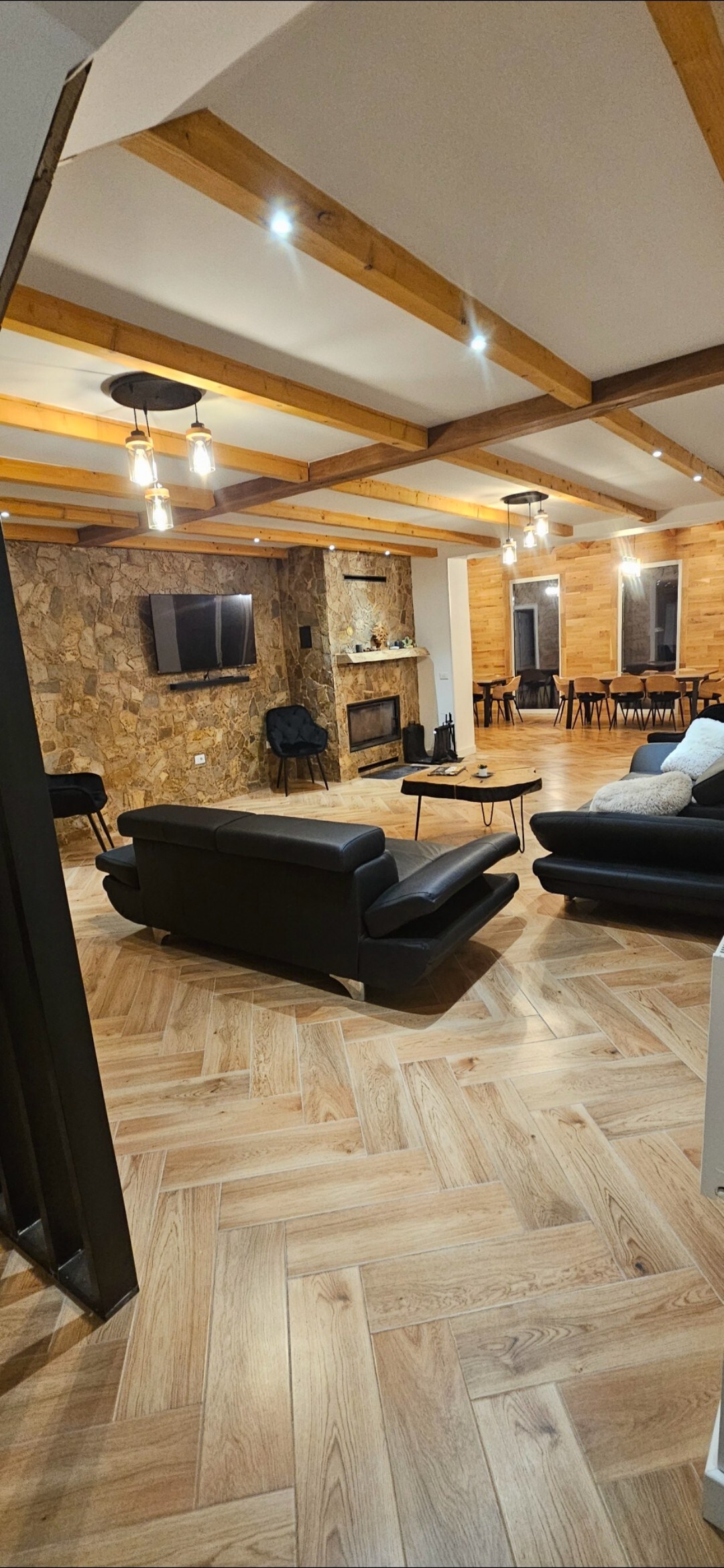 An open living area features a combination of wooden beams and stone walls. Comfortable seating arrangements include black couches around a central coffee table. A television is mounted on the wall, while warm lighting enhances the inviting atmosphere of the space.