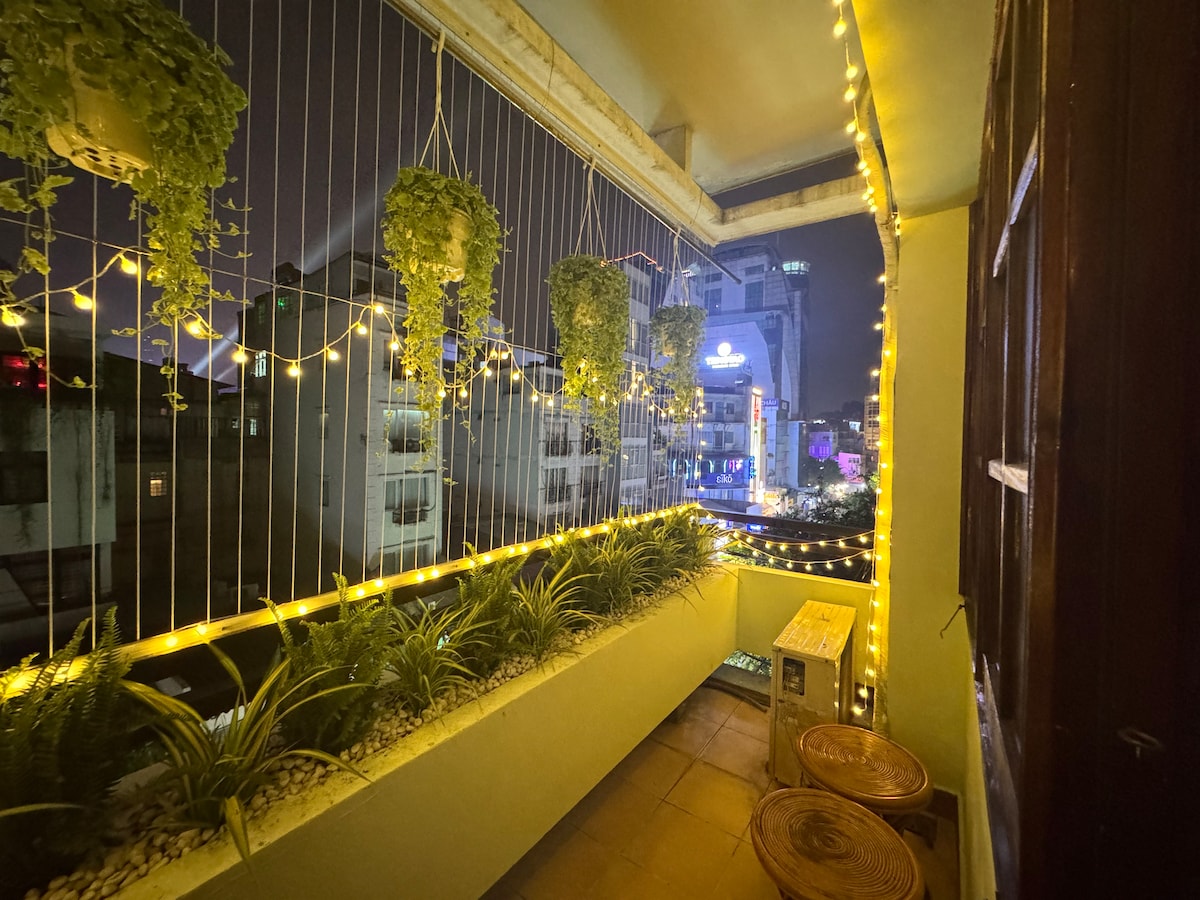 A well-lit private balcony is adorned with hanging plants and string lights, creating a cozy space for relaxation. The view showcases vibrant city lights in the background, while two wooden chairs are positioned for comfort. A small table sits against the wall, enhancing the outdoor ambiance.
