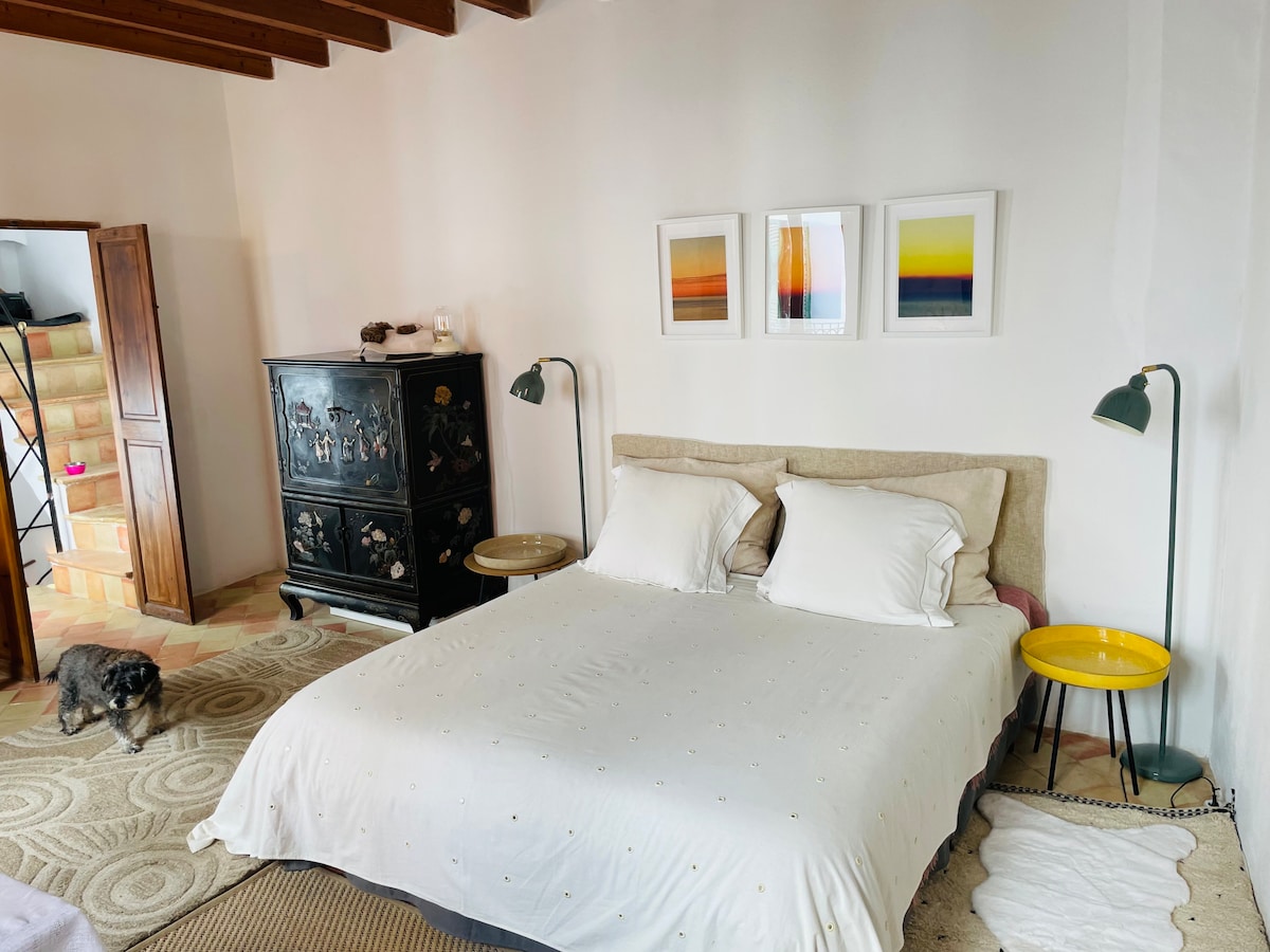 A spacious bedroom features a comfortable bed with soft linens and two unique bedside lamps on matching tables. Three framed artworks adorn the wall above the bed, contrasting with the neutral tones of the room. A small dog is seen resting on the floor.
