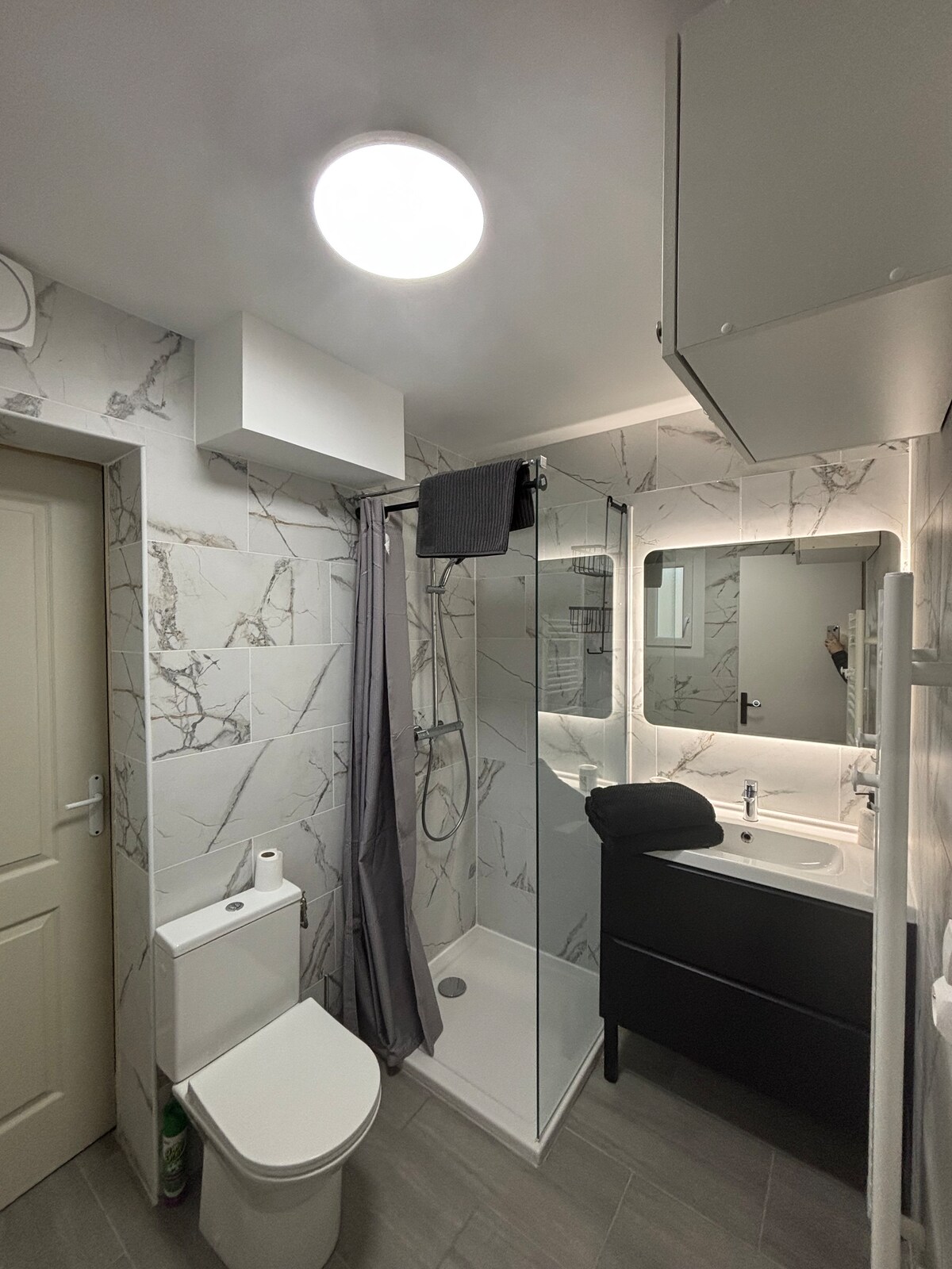 The bathroom features modern fixtures with a walk-in shower enclosed by clear glass. A sleek vanity is accompanied by a mirror, while a compact toilet and minimalist storage are positioned conveniently. Marble-patterned walls and neutral tones create an elegant, clean space.