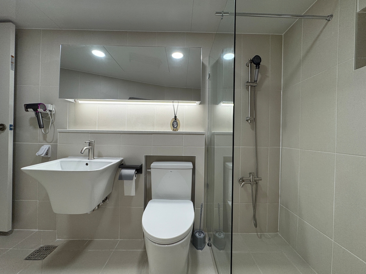 A spacious bathroom is featured with a modern toilet and a wall-mounted sink. A large mirror reflects the well-lit area, with shower fixtures positioned behind a glass enclosure. Neutral tiles line the floor and walls, contributing to a clean aesthetic.