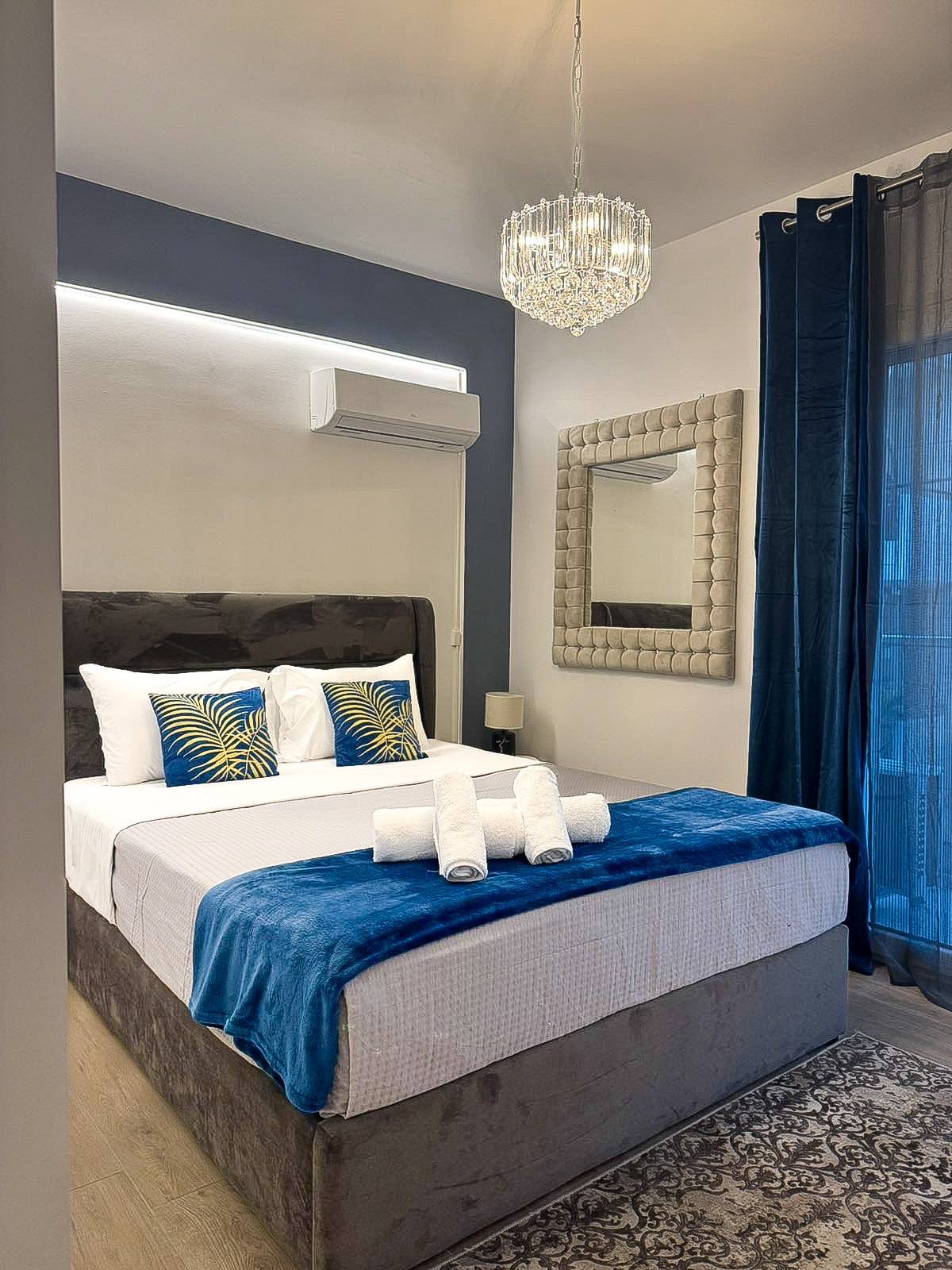 A well-appointed sleeping area features a cozy bed with decorative pillows and a soft blanket. An elegant chandelier hangs above, and a large mirror enhances the space. Dark curtains provide privacy, and natural light filters in from a nearby window.