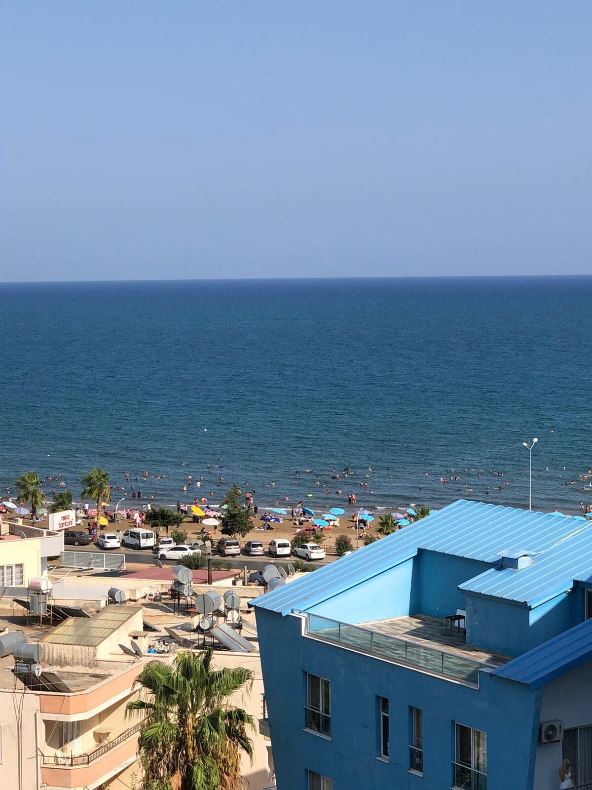 A panoramic view of the coastline stretches across the horizon, revealing a vibrant beach below. Sunbathers can be seen enjoying the shore as colorful umbrellas dot the sandy area. The clear blue sea meets a cloudless sky, enhancing the serene atmosphere.