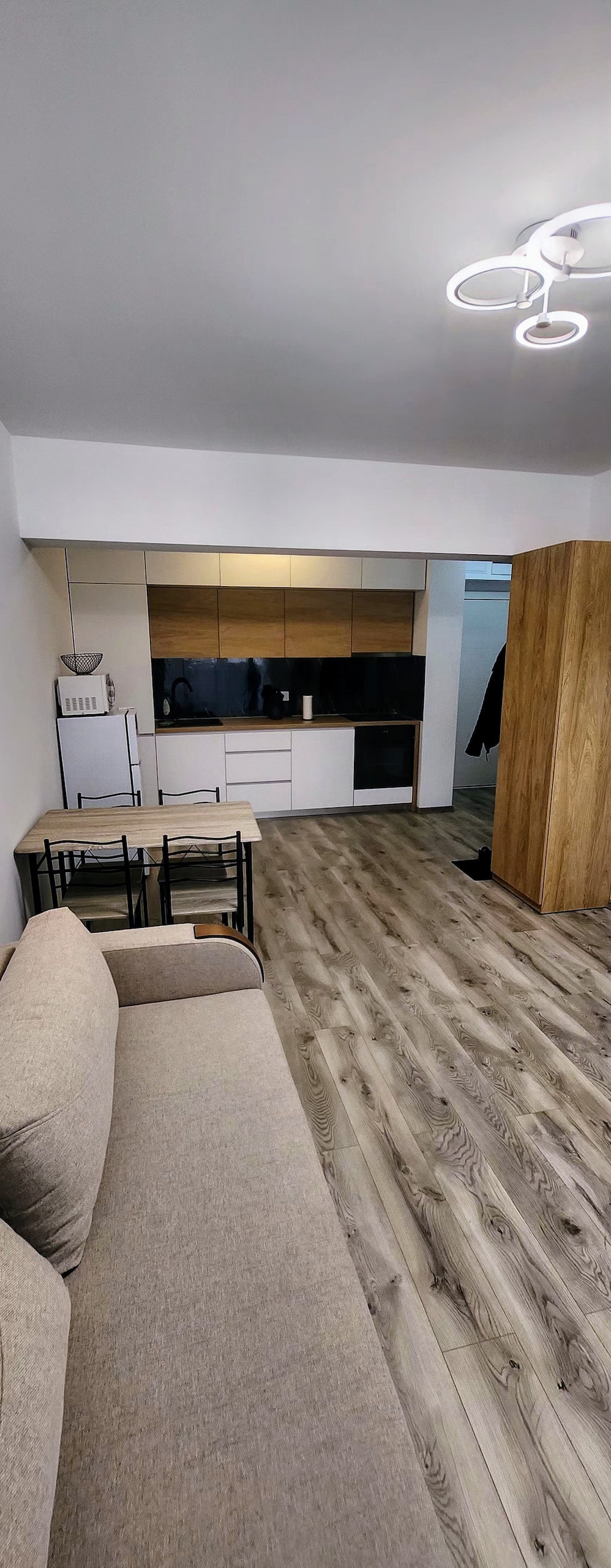 A modern living space features a light-colored sofa placed near a small dining table with black chairs. A kitchenette with dark cabinetry and appliances is visible in the background, while warm wood flooring enhances the room's aesthetic.
