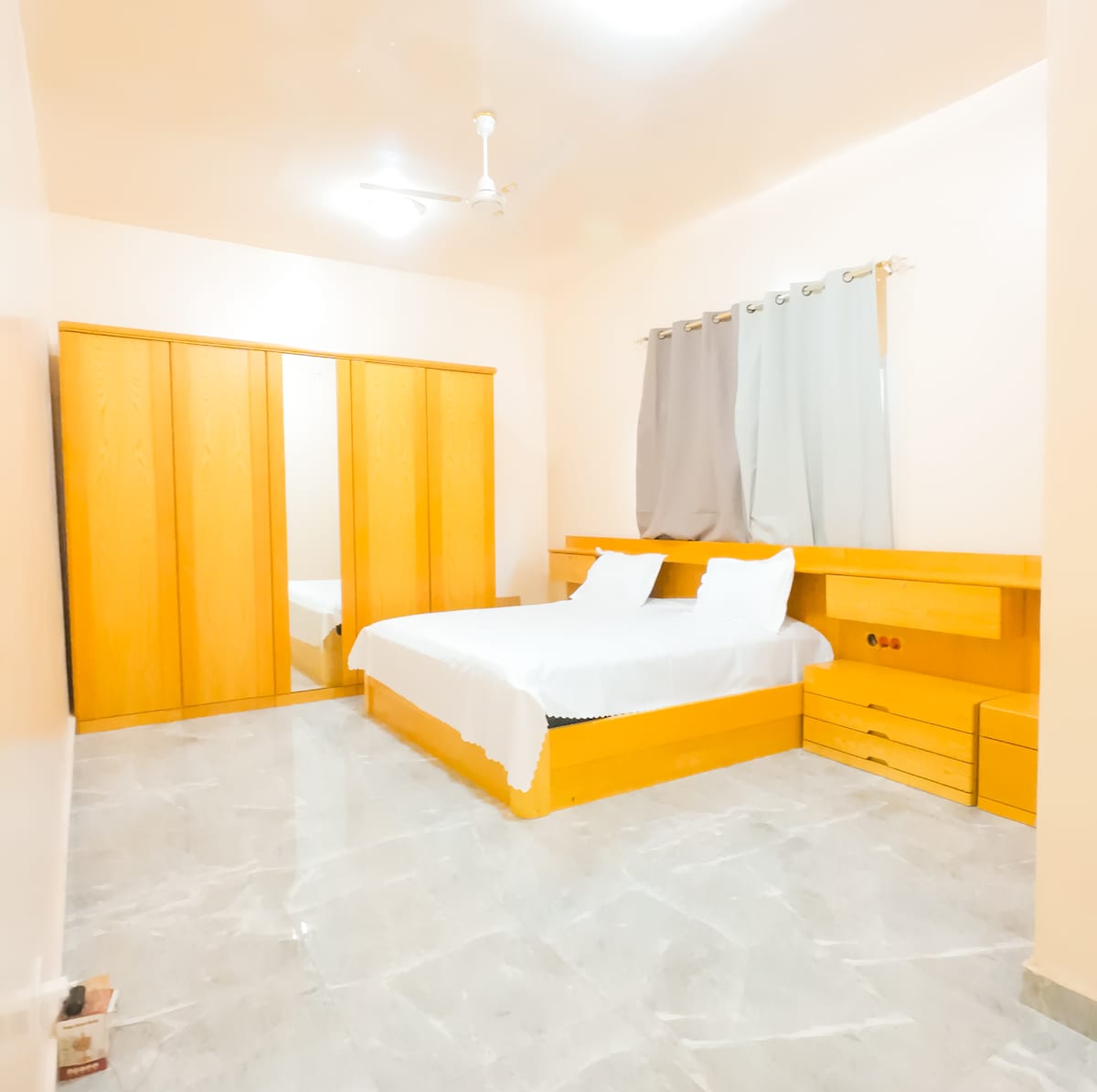 A spacious bedroom features a large bed with white linens, complemented by a wooden headboard and bedside storage. Natural light is allowed in through curtains on the window, and a mirrored wardrobe adds functionality to the room's design.
