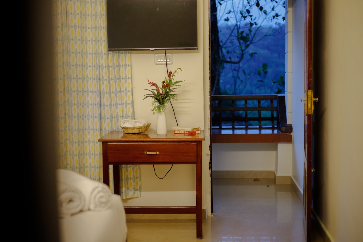 A simple wooden table is positioned next to a bed, adorned with a small vase containing fresh flowers. A television is mounted on the wall opposite the bed. Light from a nearby window highlights a view of greenery in the background.