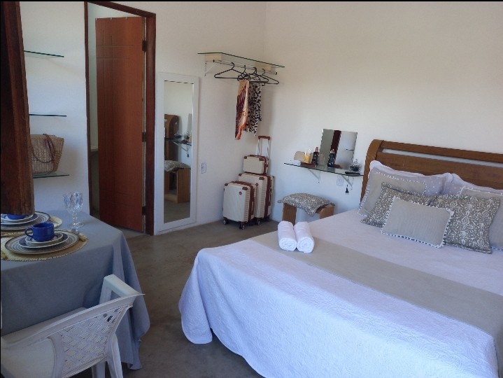 A bright and airy suite features a spacious bed with decorative pillows. A small dining table is set nearby, complemented by elegant dishware. Storage solutions include a rack with clothes and a mirror. Natural light fills the space, enhancing its open feel.