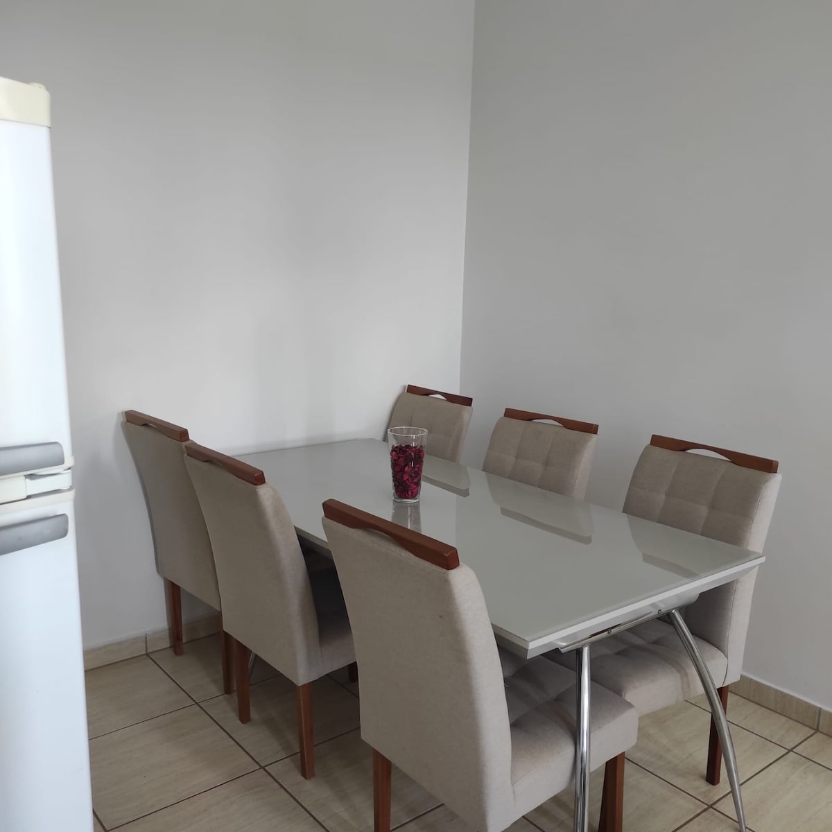 A clean dining area features a glass table surrounded by six upholstered chairs. A decorative bowl filled with fruit sits at the center of the table. The walls are painted in a light color, and the area is well-lit, creating a welcoming environment.