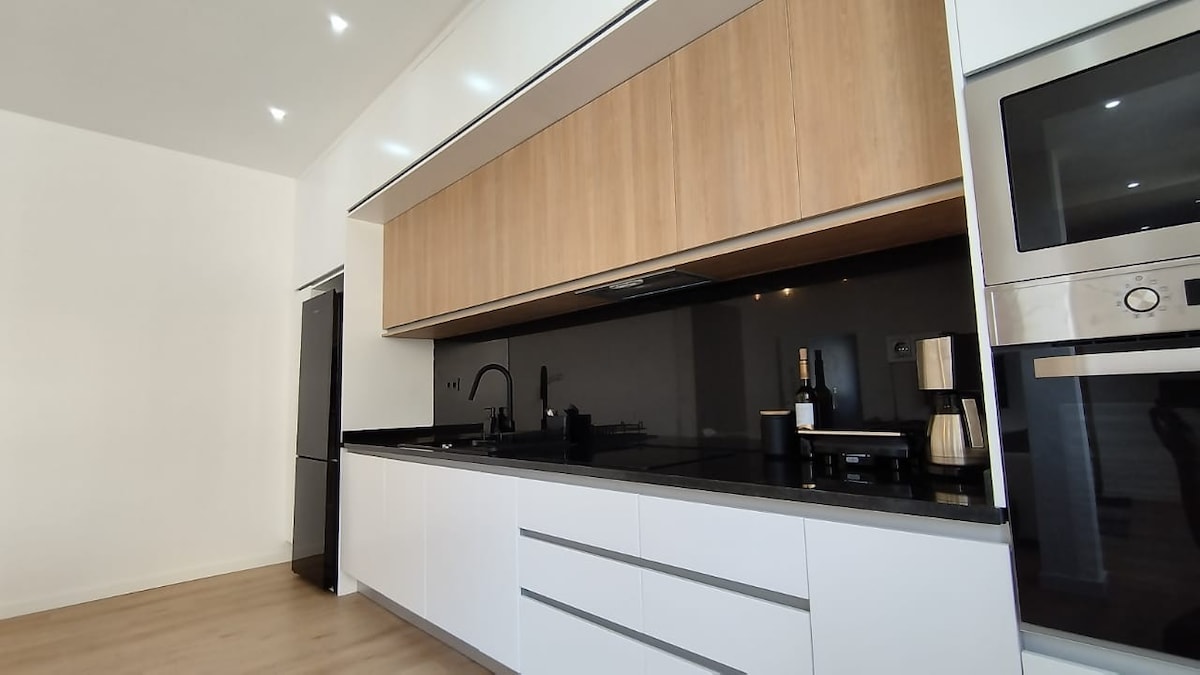 A modern kitchen is highlighted by sleek cabinetry featuring a wood and white finish. Black countertops complement the minimalist design, while appliances, including a coffee maker and a refrigerator, are neatly arranged. The stylish layout promotes functionality and a clean aesthetic.