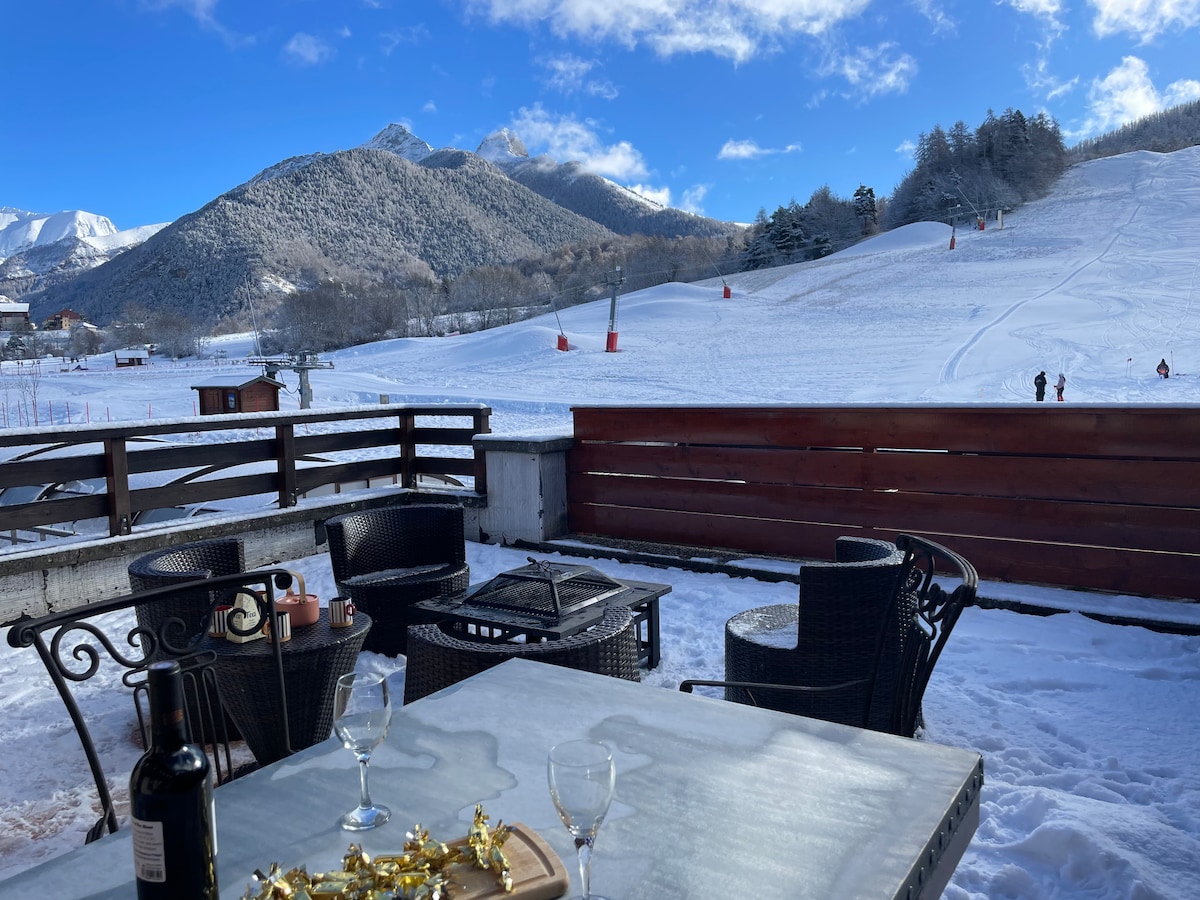 A terrace area is visible, furnished with a table and several chairs. The snow-covered slopes and distant mountains create a scenic backdrop, with ski lifts in view. Trees line the horizon under a clear blue sky, enhancing the outdoor atmosphere.