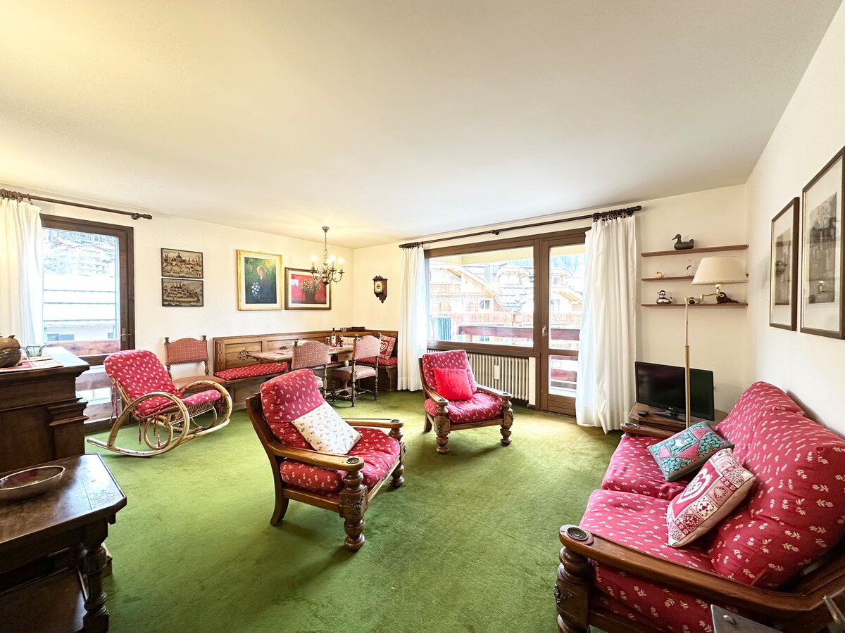 A spacious living area is highlighted by a soft green carpet and features rustic-style furnishings, including two red-upholstered armchairs and a matching couch. A dining table for six is positioned in the background, adjacent to large windows that provide a view of the surrounding landscape.