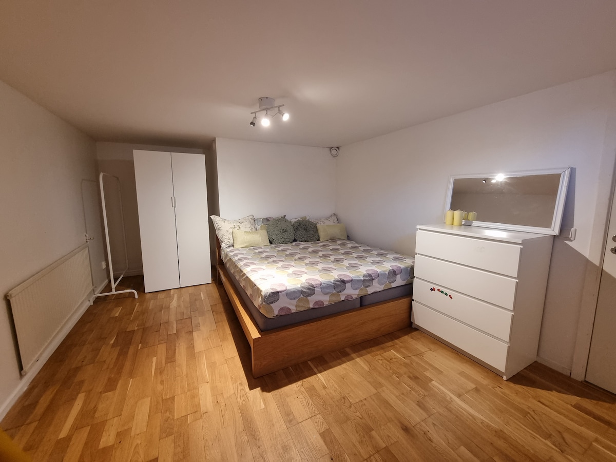 A spacious sleeping area features a large bed with a patterned comforter and decorative pillows. A white dresser with a mirror reflects the room's light. A wardrobe and a clothing rack provide storage, while warm wooden flooring adds to the inviting atmosphere.