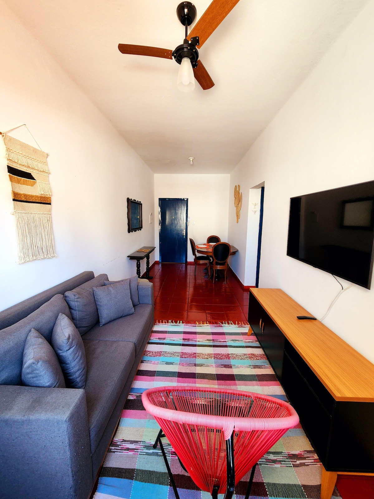 A bright living space is featured with a gray sectional sofa positioned against a white wall, complemented by colorful accent pillows. A traditional rug adds texture to the red tiled floor. A wall-mounted TV and a wooden sideboard are also visible, with a circular chair providing additional seating.
