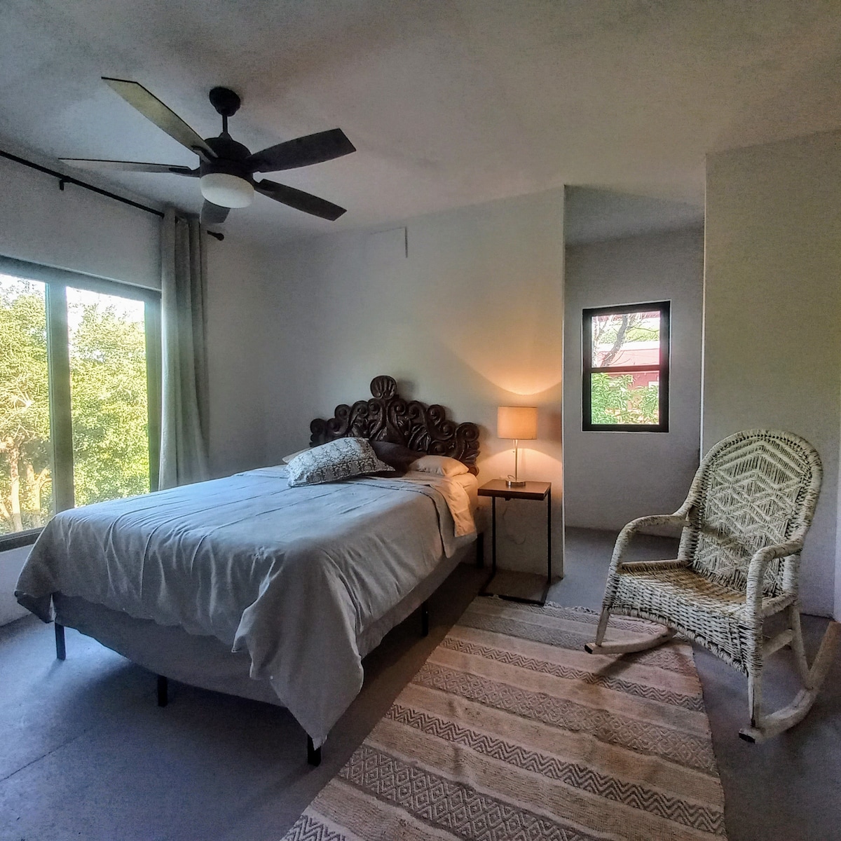 A spacious bedroom is featured, furnished with a large bed and decorative headboard. Natural light streams in through large windows, enhancing the serene atmosphere. A bedside lamp and a comfortable rocking chair add to the cozy ambiance, while a soft area rug lies beneath.