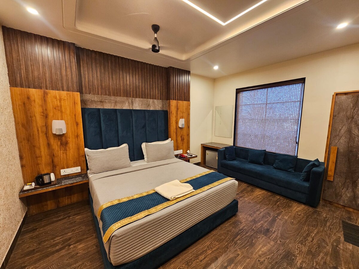 A modern guest room features a spacious bed with a decorative bed runner. A seating area with a plush couch is situated against the wall. Soft lighting highlights the wooden accents and window treatments, and a work desk is positioned nearby.