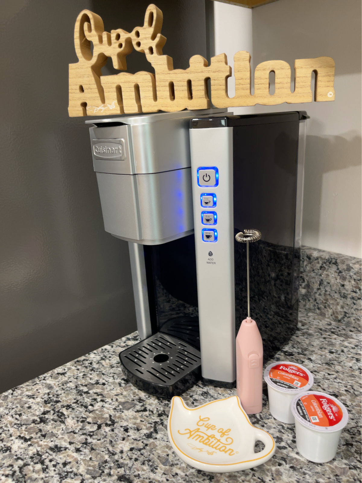 A modern coffee maker is displayed on a speckled granite countertop. Next to it, a decorative sign reading 'Cup of Ambition' is placed above a small plate that holds coffee capsules. A frother lies nearby, enhancing the coffee preparation setup.