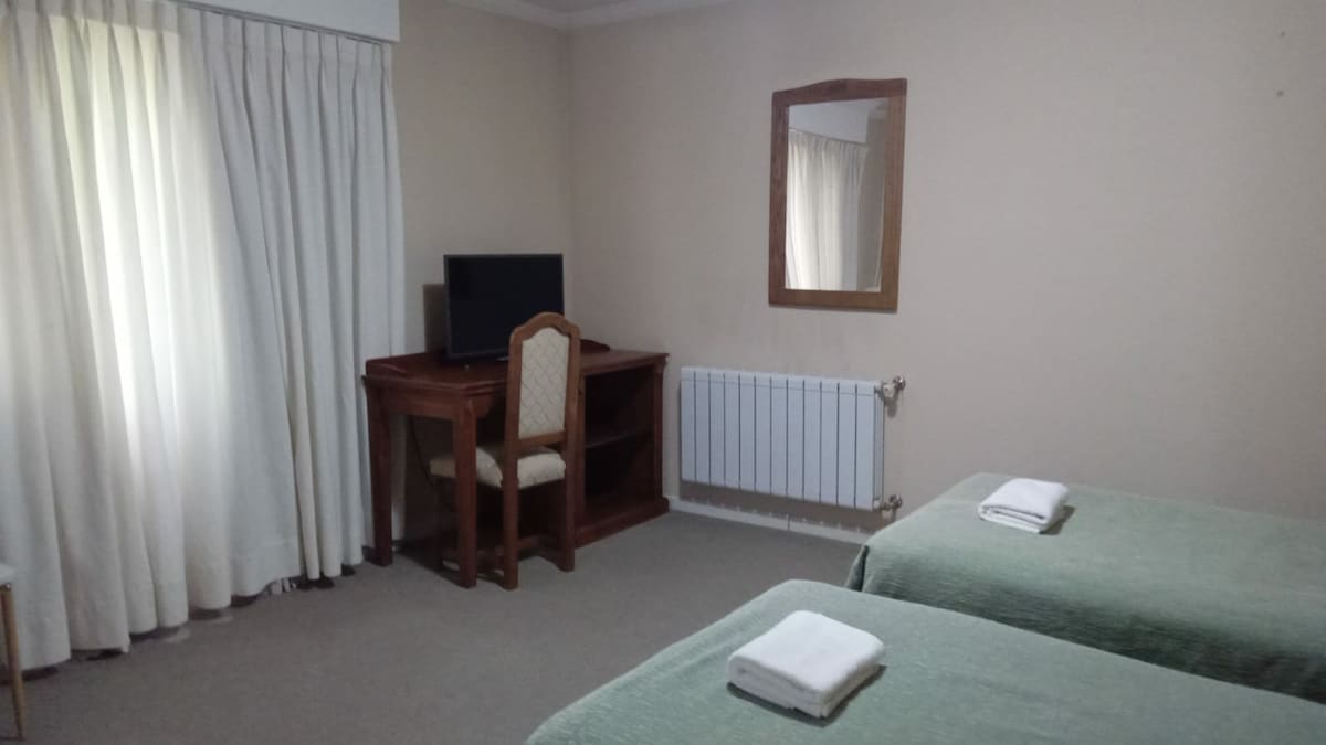 The room features two beds dressed in soft green blankets. A wooden desk with a chair is positioned next to a wall-mounted television. Natural light enters through a nearby window adorned with white curtains, while a simple mirror is mounted above the desk.