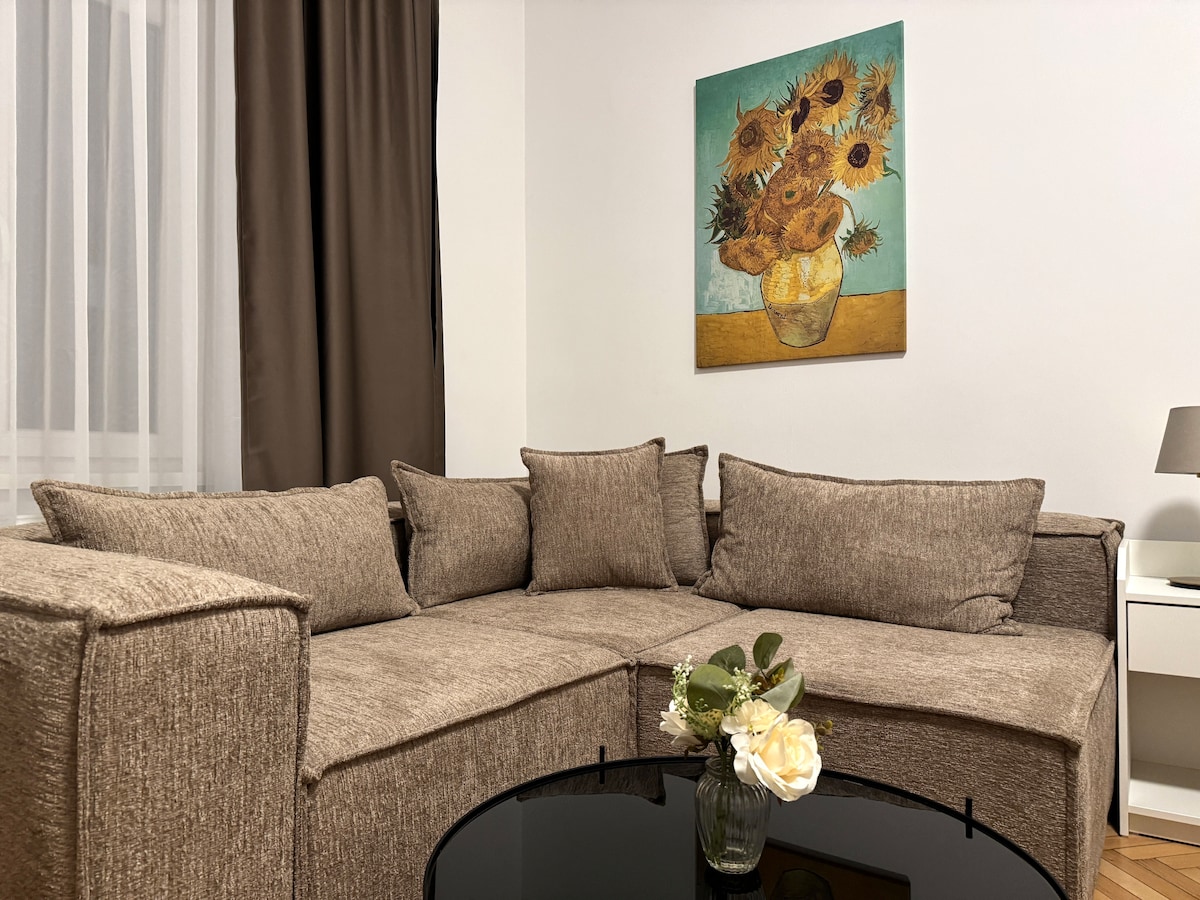 A comfortable sectional couch in a neutral tone is positioned in a cozy corner of the room. A vibrant artwork featuring sunflowers hangs on the wall above it. A round black coffee table is set in front, adorned with a small vase of white flowers.