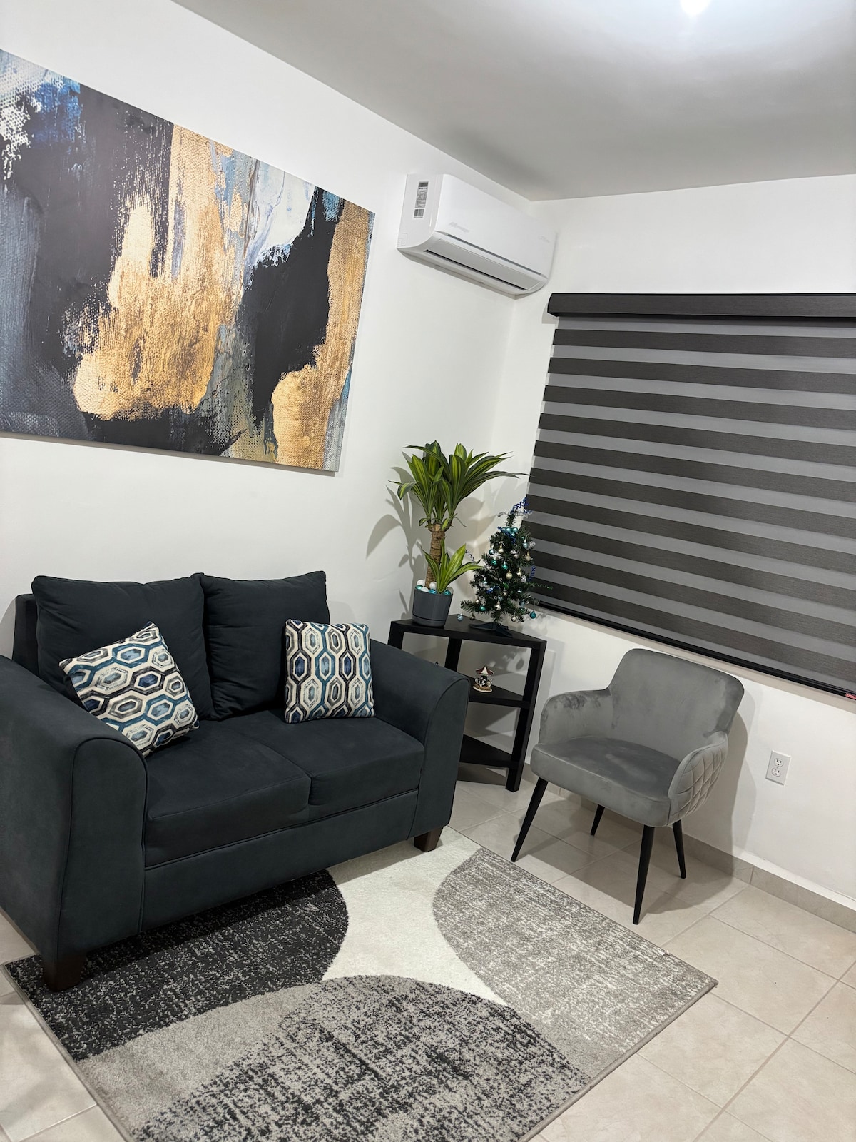 A cozy seating area features a dark grey sofa with decorative cushions, accompanied by a minimalist grey chair. A stylish area rug adds pattern to the tiled floor, while a potted plant enhances the space. Soft natural light filters through a modern striped window treatment.