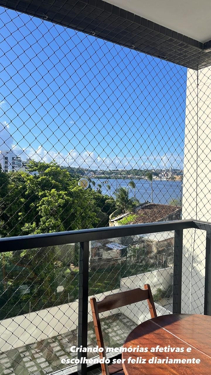A balcony view presents a serene landscape featuring a river surrounded by lush greenery. The clear blue sky contrasts with the buildings in the background, creating a calm and inviting outdoor space. A wooden table is positioned in the foreground, ideal for enjoying the view.
