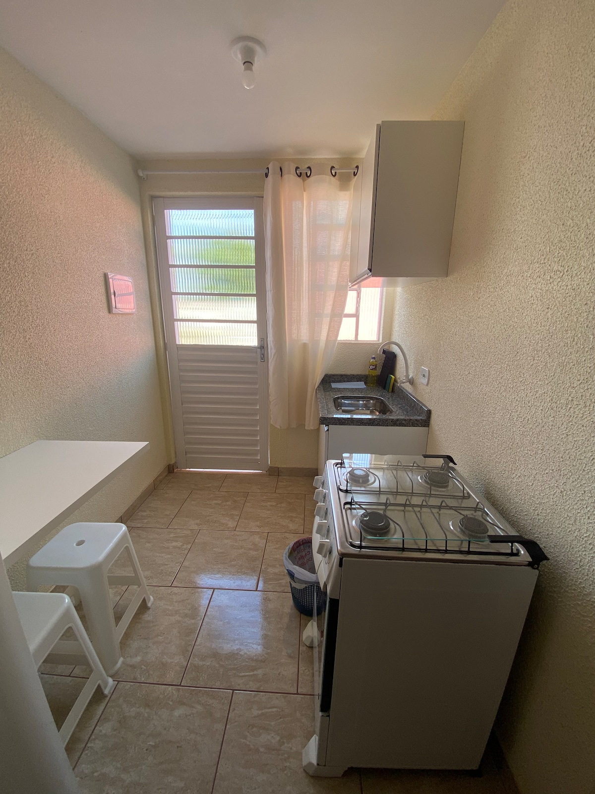 A compact kitchen area features a gas stove alongside a sink and counter space. A small table with two stools is positioned near a window, allowing natural light to brighten the light-colored walls. A door leads to the outside, fitted with a windowed panel.