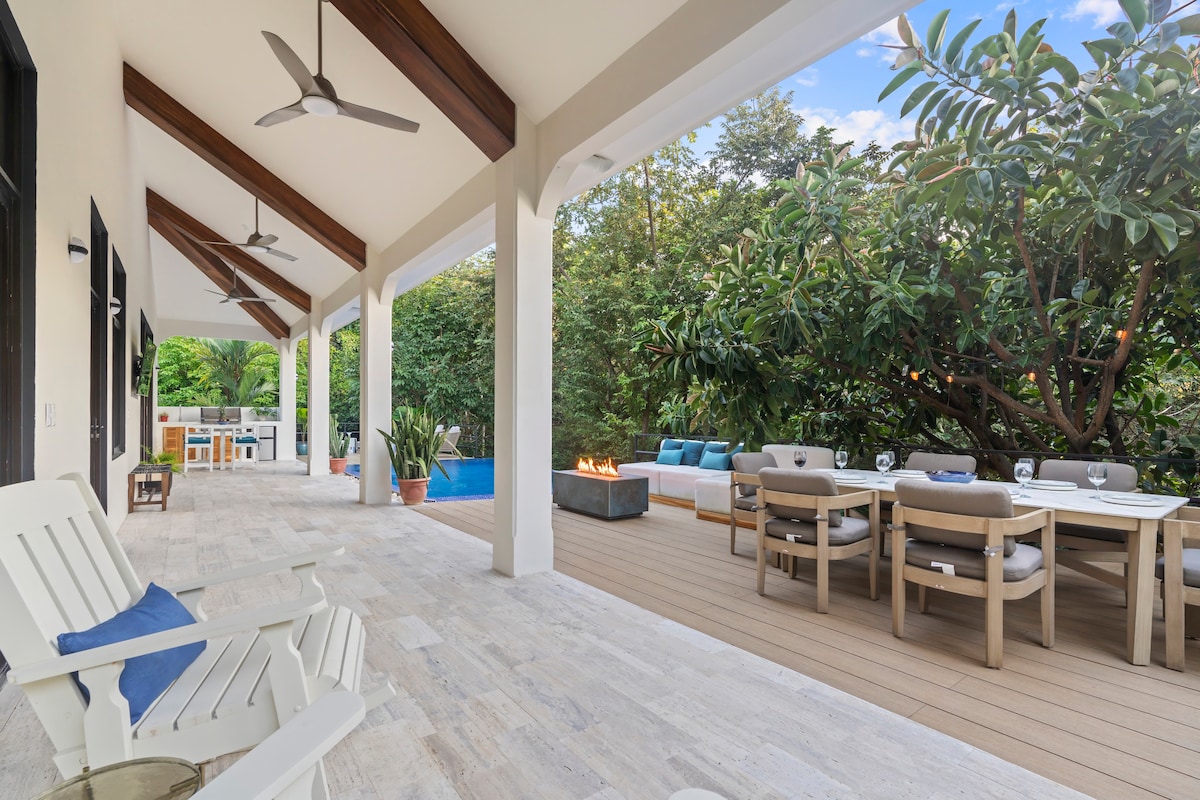 A spacious outdoor patio features comfortable seating and a dining area, surrounded by lush greenery. The private pool and barbecue area are visible in the background, with a fire feature adding warmth to the inviting space.