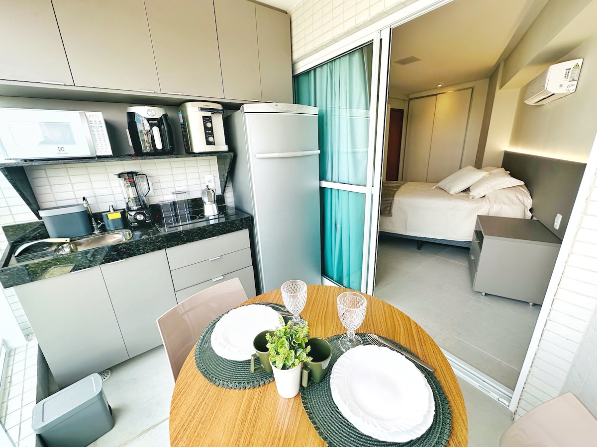 A modern kitchen area features sleek cabinets and a dark granite countertop. A dining table, set for two, is adorned with plates and a small plant. The adjoining bedroom is visible through a glass door, offering a glimpse of a spacious bed with neatly arranged pillows.