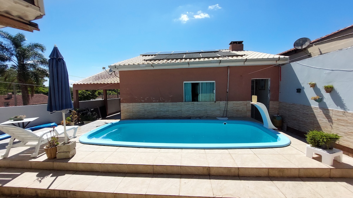 A sunlit backyard features a swimming pool surrounded by tiled flooring. A lounge area with a blue umbrella and deck chairs is positioned beside the pool. The house in the background displays a warm color exterior with a patio roof and several windows.