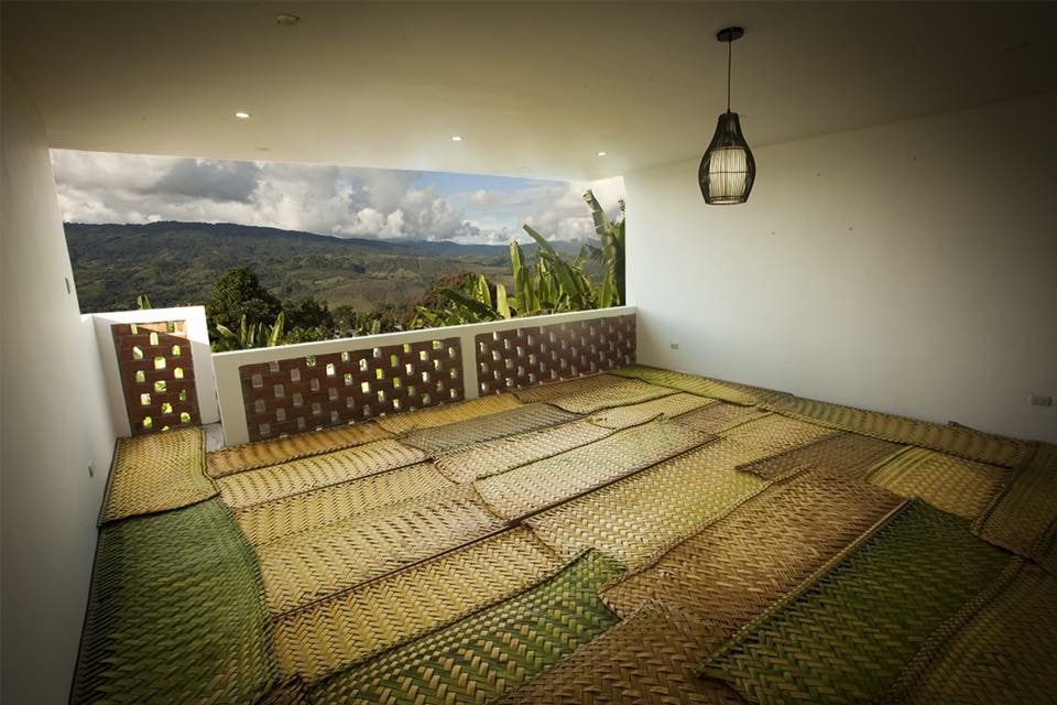 An open terrace area is furnished with colorful woven mats, providing a comfortable space for relaxation or meditation. The expansive view of the lush landscape can be seen through the decorative walls, enhancing the serene outdoor ambiance.