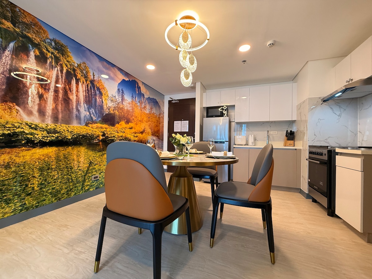 A dining area showcases a round table with a golden base surrounded by four modern chairs. A mural of a serene landscape with waterfalls and autumn foliage adorns the wall. The kitchen area features sleek white cabinetry and modern appliances, creating a cohesive design.