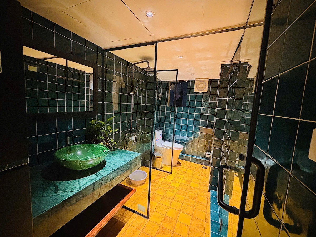 A spacious bathroom features dark green tiled walls and a bright yellow tiled floor. A large bowl sink rests on a wooden countertop, complemented by a framed mirror. A glass shower enclosure is visible, alongside a toilet positioned within the well-lit area.