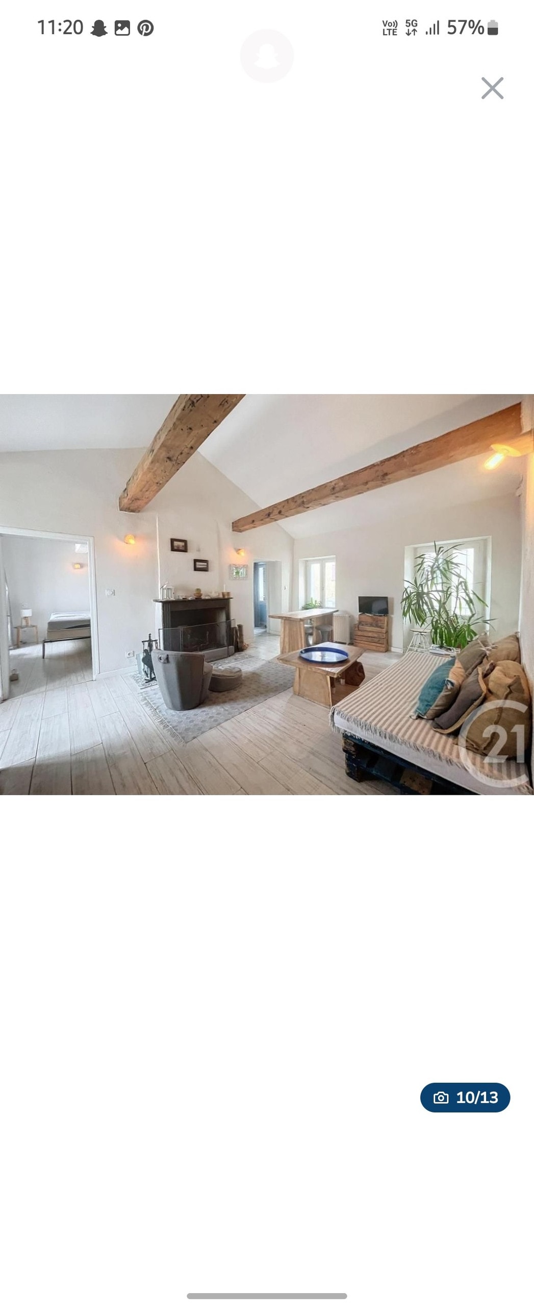 A light-filled living area features exposed wooden beams and a functional fireplace. A cozy seating area is positioned in front of the fireplace, with large windows allowing natural light to brighten the space. The room connects to the adjacent kitchen area, visible in the background.