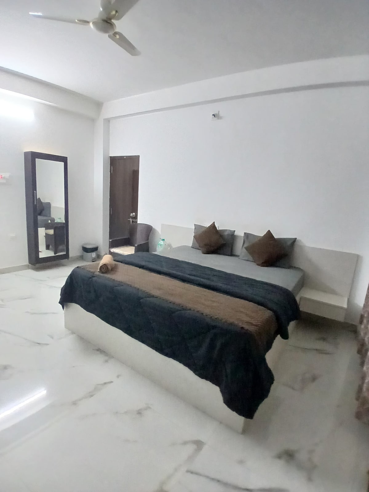 A spacious bedroom is furnished with a large bed featuring neutral-toned linens and pillows. A full-length mirror is positioned beside the bed, reflecting the minimalist decor. A chair is placed nearby, with a soft light illuminating the room from a single ceiling fixture.